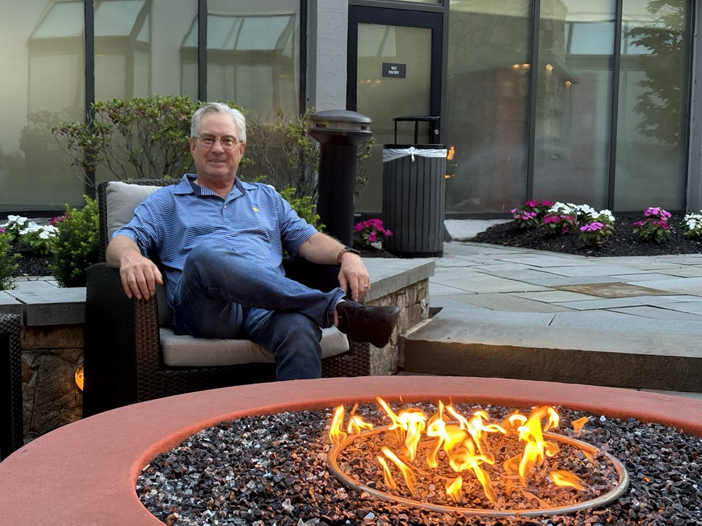 Craig Spiesman seated with crossed legs near an outdoor fire feature and garden flowers
