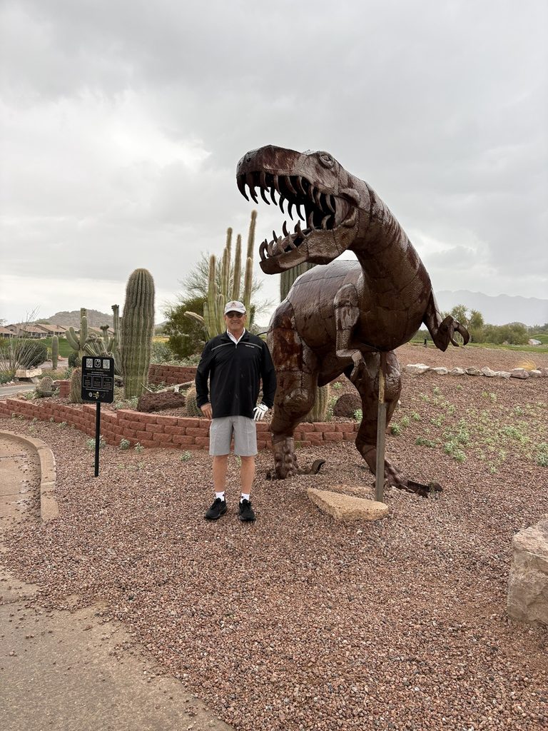 Craig Spiesman standing in a desert garden beside a large metal dinosaur sculpture
