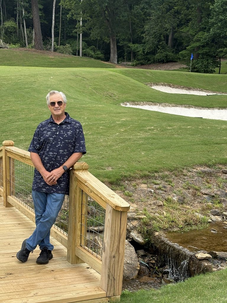 Craig Spiesman standing outdoors with a golf flag, bunkers, and woodland in background
